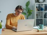 Man in yellow sweater working on laptop at desk.